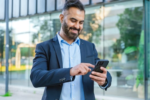 Smiling middle-aged businessman in a blue shirt holding and using a smartphone outdoors in a modern urban setting.