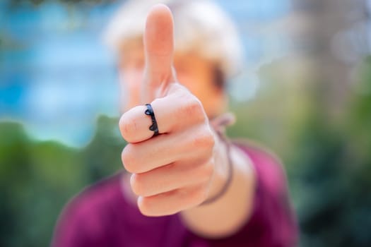 Close-up of a man giving a thumbs-up gesture, wearing a black ring on his thumb. The background is blurred, with greenery and urban elements visible.