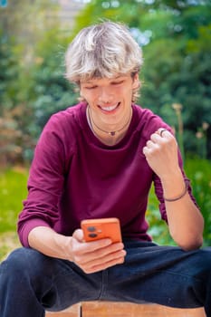 Smiling young man in a burgundy shirt sitting outdoors, looking at his smartphone with a fist pump gesture of celebration, surrounded by greenery