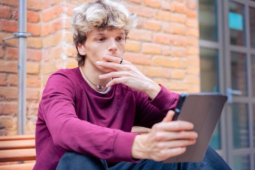 Young man in a burgundy shirt sitting against a brick wall, holding a tablet and looking thoughtful, outdoors in an urban setting