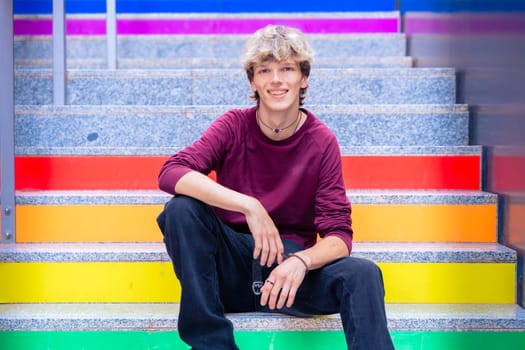 Young man in a burgundy shirt sitting casually on rainbow-colored stairs, smiling confidently, with a modern urban backdrop.