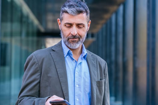 Smiling middle-aged businessman in a blue suit holding and using a smartphone outdoors in a modern urban setting.