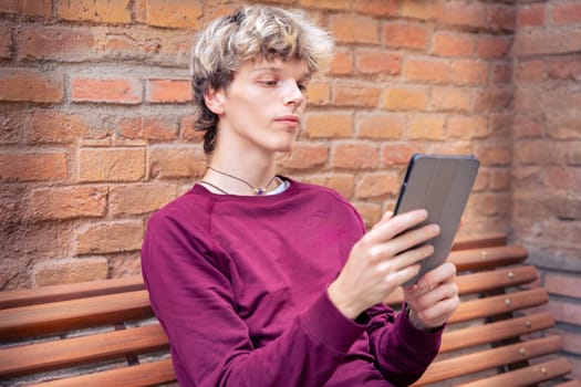 Young man in a burgundy shirt sitting against a brick wall, holding a tablet and looking thoughtful, outdoors in an urban setting