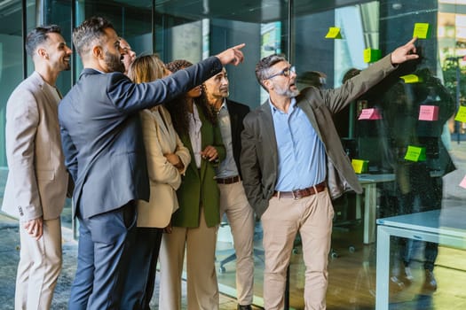 A diverse group of business professionals brainstorming in a modern office. A man points to sticky notes on a glass wall as the team collaborates with focus and engagement.