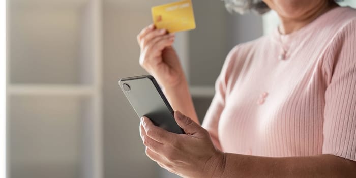 An attentive senior woman preparing for online shopping, showcasing her credit card while using her smartphone in a well lit and stylish home area.