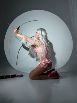 blonde girl holding bow and arrow on valentine's day. She has a big wooden bow in her hands. The girl is beautiful with bright makeup. She is wearing pink shoes. photo taken in studio