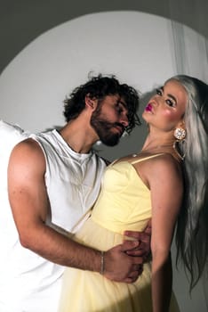 A man dressed as an angel stands next to a blonde girl. The young man has white wings. The girl is wearing a long yellow dress. They are in love and looking at each other, photo for valentine's day