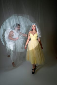 A man dressed as an angel stands next to a blonde girl. The young man has white wings. The girl is wearing a long yellow dress. They are in love and looking at each other, photo for valentine's day