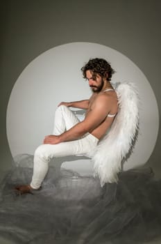 a man with white wings sits on a chair. High quality photo, a man like an angel or cupid sits on a chair and thinks. he has dark hair and a beard. the photo was taken in a studio