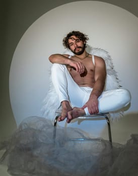 a man with white wings sits on a chair. High quality photo, a man like an angel or cupid sits on a chair and thinks. he has dark hair and a beard. the photo was taken in a studio
