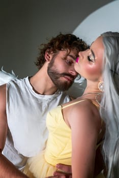 A man dressed as an angel stands next to a blonde girl. The young man has white wings. The girl is wearing a long yellow dress. They are in love and looking at each other, photo for valentine's day