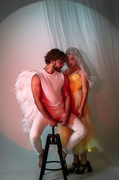 A man dressed as an angel stands next to a blonde girl. The young man has white wings. The girl is wearing a long yellow dress. They are in love and looking at each other, photo for valentine's day
