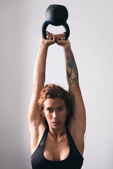 concentrate strong woman bodybuilder with tattoos lifting heavy kettlebell while training in gym against white wall, concept of fitness and active lifestyle