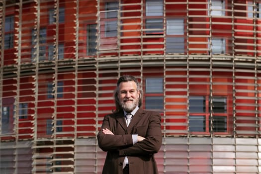 positive middle age male with beard in elegant suit standing with arms crossed and looking at camera against urban building, concept of business and mature man lifestyle, copyspace for text