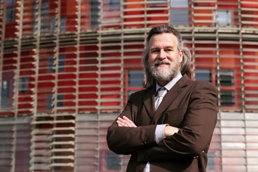 positive middle age male with beard in elegant suit standing with arms crossed and looking at camera against urban building, concept of business and mature man lifestyle, copyspace for text