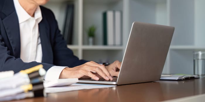 An elderly businessman concentrating on his laptop, engaged in work tasks amidst an organized workspace with documents and greenery.