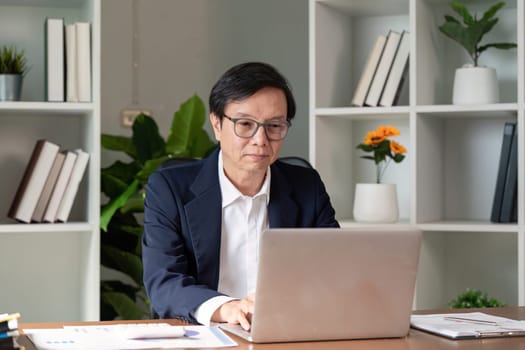 A senior professional wearing glasses diligently working on a laptop, analyzing reports in a well organized modern office setting, surrounded by greenery.