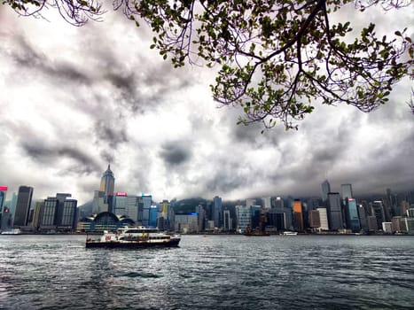 Hong Kong - April 06, 2024: A dense fog envelops the skyline of Hong Kong as clouds loom overhead, casting a gray hue over Victoria Bay. A colorful ferry navigates the water, adding life to the misty atmosphere.