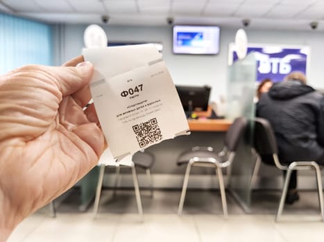 Kirov, Russia - October 08, 2024: A hand holds a ticket with the queue number 047 at a bank, while customers wait at the counter.