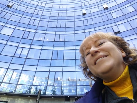 This cheerful middle aged woman takes a selfie against a backdrop of a modern glass building while enjoying her outing.