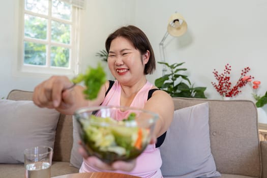 A joyful overweight woman presents a bowl of fresh salad with a big smile, showcasing her enthusiasm for healthy eating and her weight loss journey in a bright, inviting home.