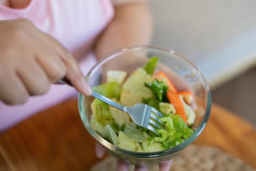 An overweight woman's hand holding a fork dips into a colorful salad, emphasizing her commitment to a healthier lifestyle and weight loss at home.