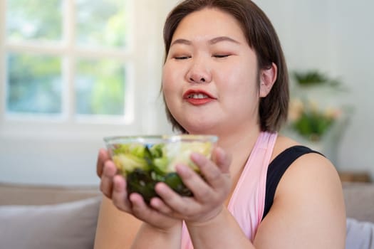 An overweight woman smiles as she holds a vibrant salad against her chest, showcasing her dedication to a healthier lifestyle and weight loss at home.