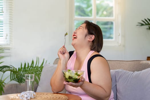 A happy overweight woman savoring a salad in her cozy living room, celebrating her weight loss journey with a smile as sunlight streams in through the window.