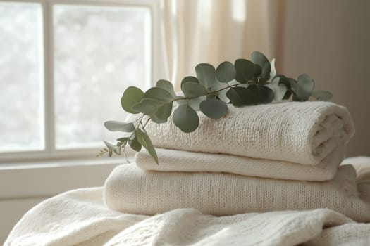 A stack of soft beige blankets placed neatly by a large window. Delicate eucalyptus branches rest on the top blanket, enhancing the serene atmosphere of the space.