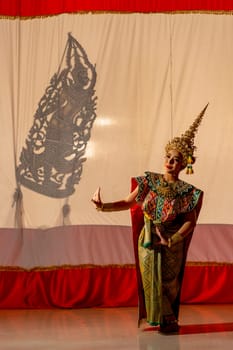 Vertical image of one woman with Khon or Mask dance dress stand with performance action in front of grand shadow play in the background with white screen.