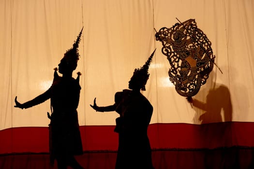 Silhouette and wide shot of man and woman with Khon or Mask dance costume stand with performance action in front of grand shadow play in the background with white screen.