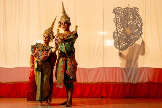 Two people of man and woman with Khon or Mask dance dress stand with performance action in front of grand shadow play in the background with white screen.