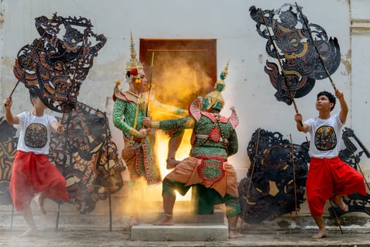 Two men with Khon mask charactor as angel and giant stand and performance action as figthing in front of door frame of old building also two men hold grand shadow play in front and warm light in back.