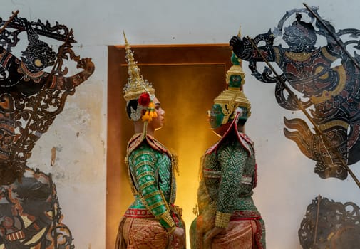 Two charactor of Khon mask costume as angel and giant stand with facing each other in front of door frame with warm light in the back.