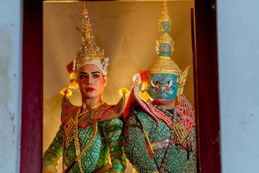 Two charactors of Khon mask costume stand with backs leaning against each other and giant mask stand in right side at door frame with beam light in the back.