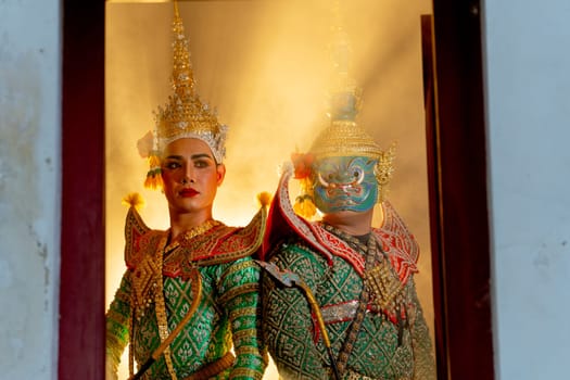 Two charactors of Khon mask costume stand with backs leaning against each other and human charactor stand in left side at door frame with beam light in the back.