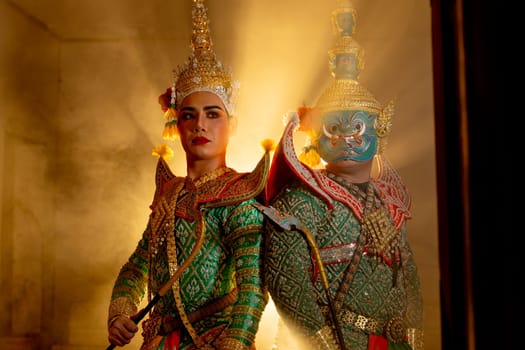 Wide shot of two charactors of Khon mask costume stand with backs leaning against each other and giant mask stand in right side at door frame with beam light in the back.