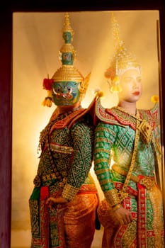 Vertical image of two charactors of Khon mask costume stand with backs leaning against each other at door frame with beam light in the back.
