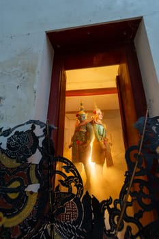 Vertical image and wide shot of two charactor of Khon mask costume stand with backs leaning against each other at door frame with light in the back and grand shadow play crafts in the front.