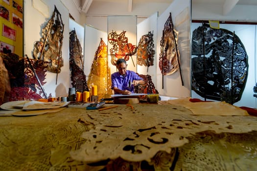Wide shot of Asian senior man wear glasses use hammer and chisel to create grand shadow play craft and he concentrate to work in area of workplace with several products are place around workplace area