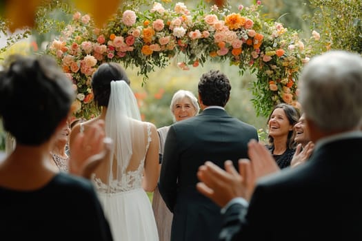 A couple stands at a floral arch amidst greenery, exchanging vows in an outdoor ceremony while guests celebrate joyfully around them.