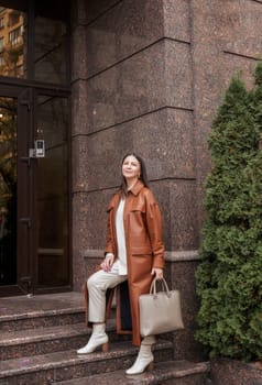 elegant woman over 40 exiting building with laptop bag. She is wearing a brown coat, holding a laptop bag. She is elegant in white shoes. She is a slim, well-groomed woman working in an office.