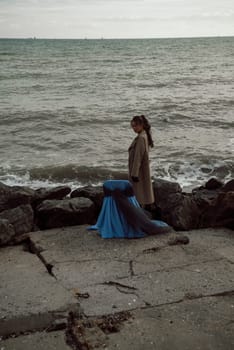 young girl on the seashore around the rocks. she is dressed in a beige trench coat, she is dreaming on the seashore among the flowers of the chrysanthemum variety. a chair decorated with blue fabric