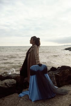 young girl on the seashore around the rocks. she is dressed in a beige trench coat, she is dreaming on the seashore among the flowers of the chrysanthemum variety. a chair decorated with blue fabric