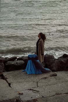 young girl on the seashore around the rocks. she is dressed in a beige trench coat, she is dreaming on the seashore among the flowers of the chrysanthemum variety. a chair decorated with blue fabric