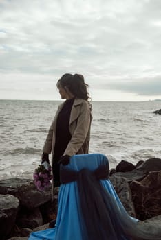 young girl on the seashore around the rocks. she is dressed in a beige trench coat, she is dreaming on the seashore among the flowers of the chrysanthemum variety. a chair decorated with blue fabric