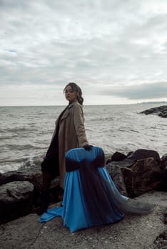 young girl on the seashore around the rocks. she is dressed in a beige trench coat, she is dreaming on the seashore among the flowers of the chrysanthemum variety. a chair decorated with blue fabric