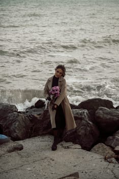 young girl on the seashore around the rocks. she is dressed in a beige trench coat, she is dreaming on the seashore among the flowers of the chrysanthemum variety. a chair decorated with blue fabric