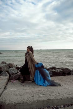 young girl on the seashore around the rocks. she is dressed in a beige trench coat, she is dreaming on the seashore among the flowers of the chrysanthemum variety. a chair decorated with blue fabric