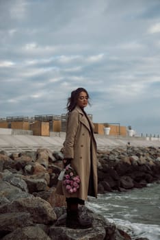 young girl on the seashore around the rocks. she is dressed in a beige trench coat, she is dreaming on the seashore among the flowers of the chrysanthemum variety. a chair decorated with blue fabric